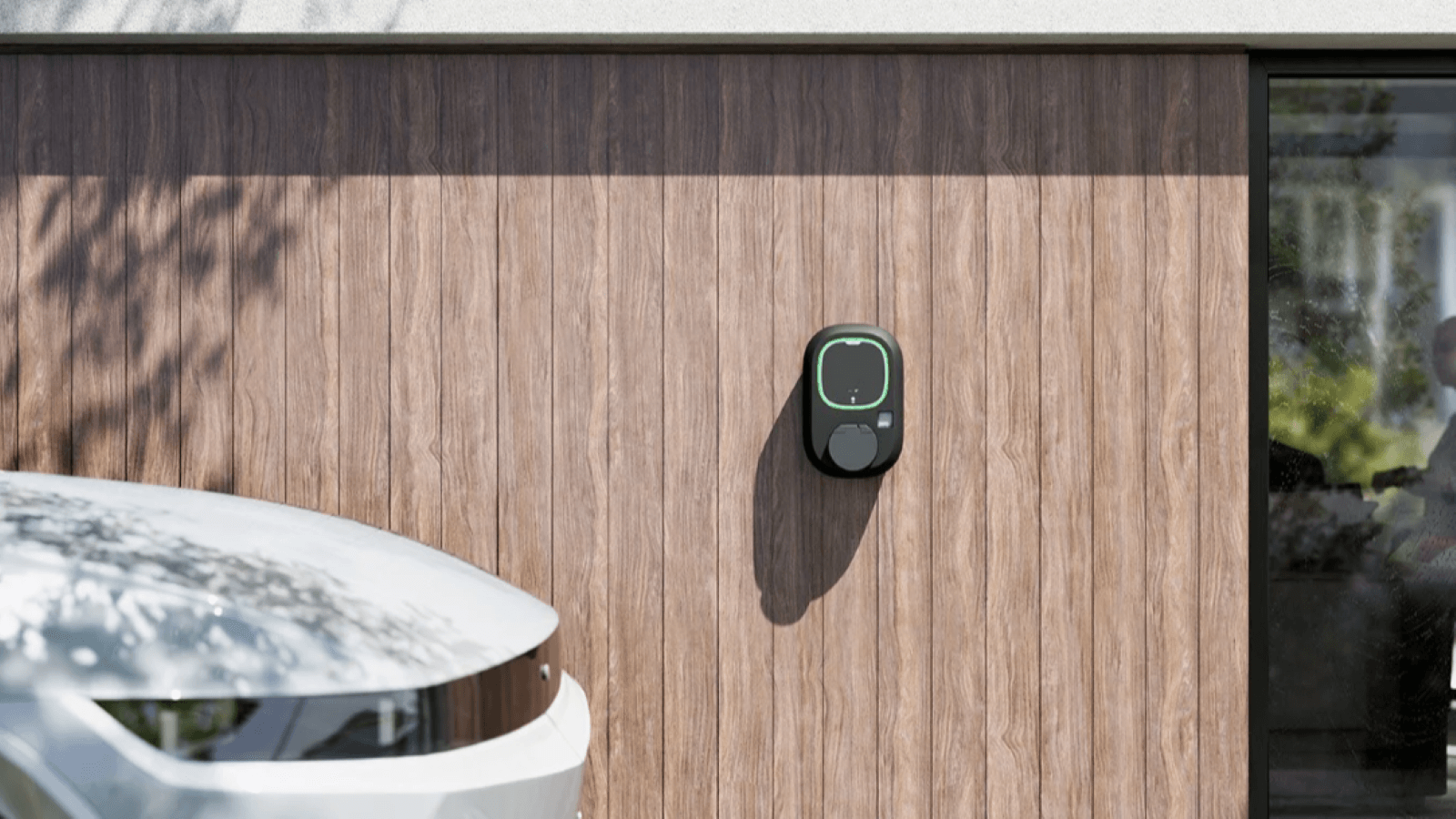 Eine kompakte Wallbox ist an einer Hauswand montiert, ein Elektroauto steht davor.