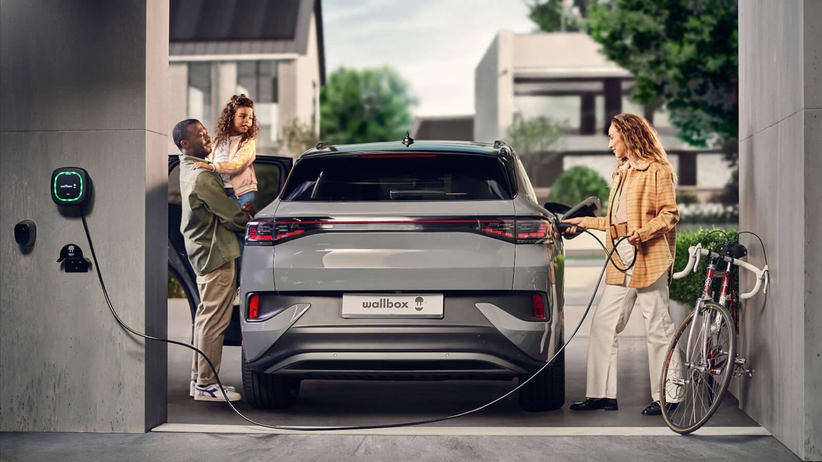 Das Bild zeigt ein Elektroauto, das in einer modernen Garage an einer Wallbox geladen wird. Eine Frau schließt das Ladekabel an, während ein Mann mit einem Kind am geöffneten Fahrzeug steht. Ein Fahrrad lehnt an der Wand.