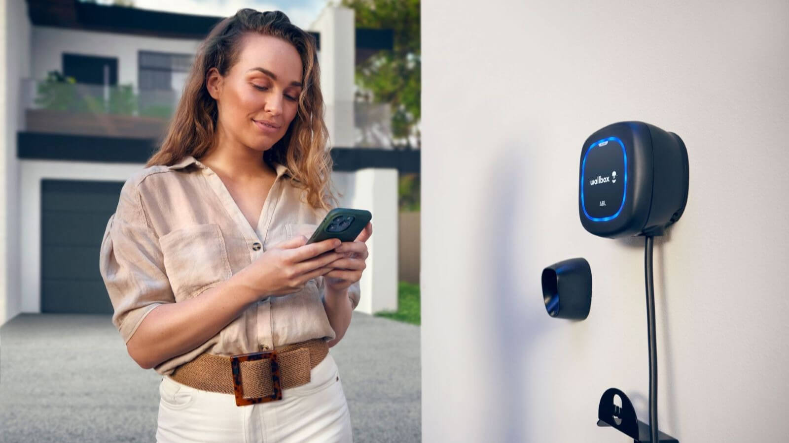 Das Bild zeigt eine Frau, die ihr Smartphone benutzt, während im Hintergrund an der Wand eine moderne Wallbox-Ladestation installiert ist.