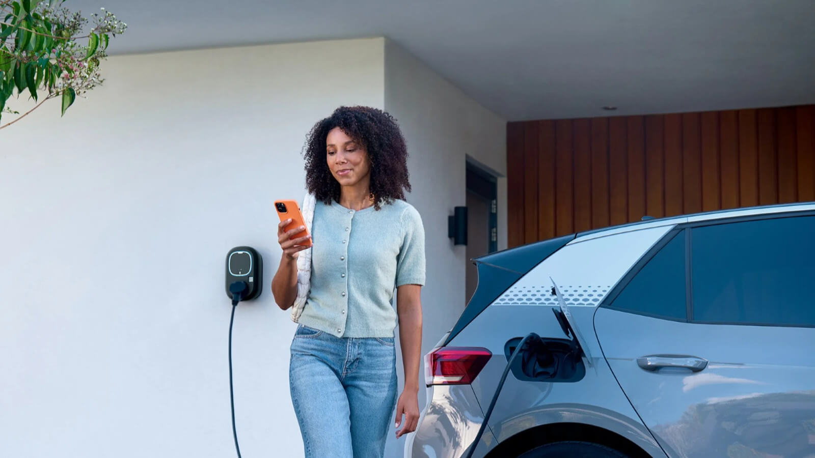 Das Bild zeigt eine Frau, die ein Smartphone in der Hand hält und an einer Wallbox-Ladestation vorbeigeht, an der ein graues Elektroauto lädt.