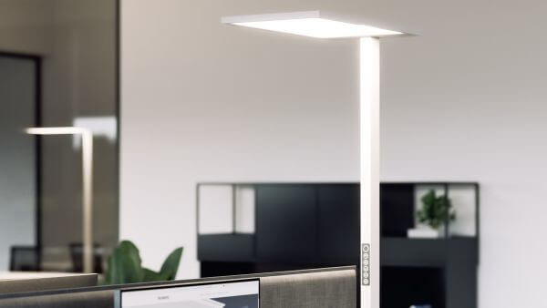 Das Bild zeigt eine moderne, weiße Stehleuchte in einem Büro, die einen Arbeitsplatz mit hellem, gleichmäßigem Licht ausleuchtet.