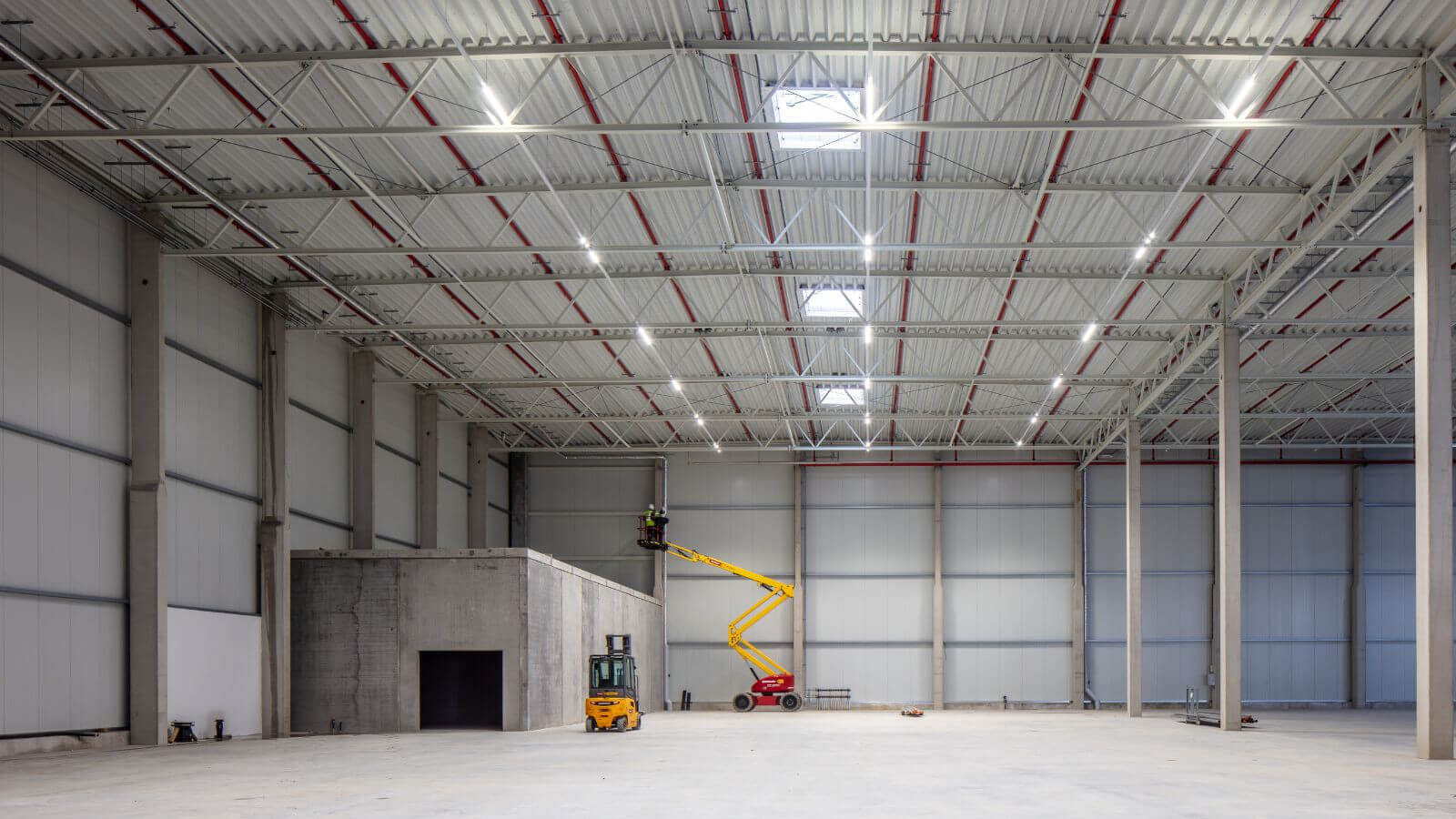 Eine große, leere Industriehalle mit hoher Decke; darin stehen ein Gabelstapler und eine Hebebühne, auf der zwei Personen an der Decke arbeiten.