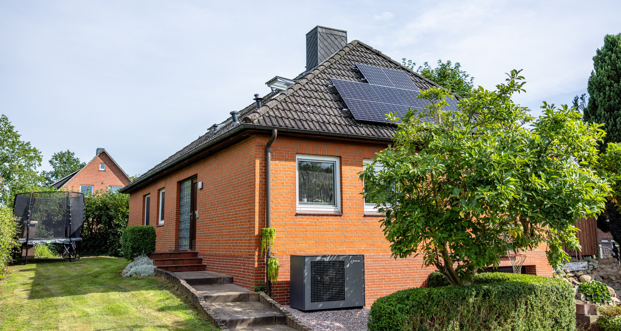 Das Bild zeigt ein Einfamilienhaus mit roter Klinkerfassade und dunkel gedecktem Satteldach. Auf dem Dach sind Solarmodule installiert. Im Vorgarten steht ein Baum und es sind gepflegte Hecken zu sehen. An der Hauswand ist eine Wärmepumpe der Marke Dimplex montiert. Im Hintergrund befindet sich ein weiteres Haus sowie ein Trampolin im Garten.