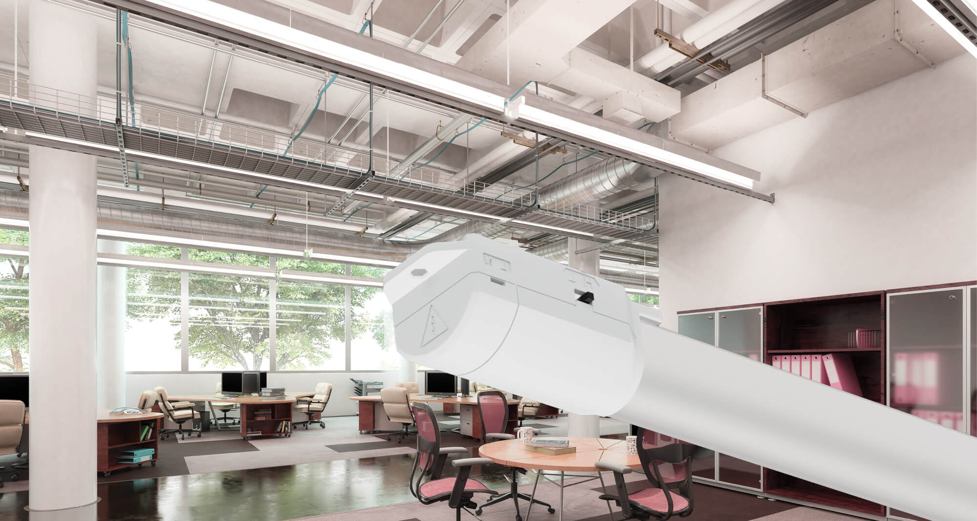 Das Bild zeigt ein modernes Großraumbüro mit mehreren Schreibtischen, Stühlen und Computern. Im Vordergrund ist eine vergrößerte Leuchte mit weißem Gehäuse zu sehen, die an der Decke montiert ist. An der Decke verlaufen außerdem sichtbare Kabeltrassen, Lüftungsrohre und weitere Installationen. Große Fenster lassen Tageslicht in den Raum.