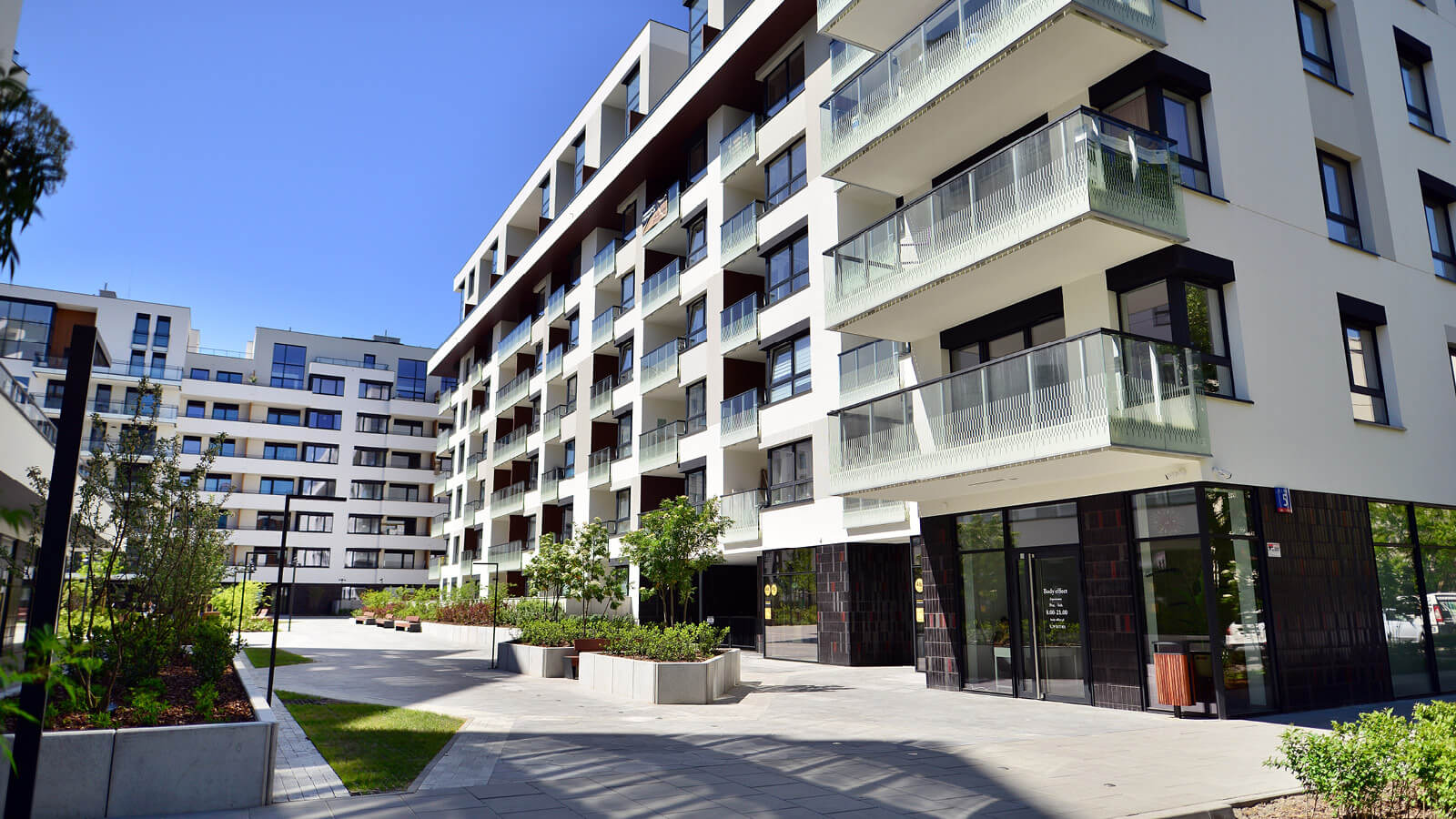 Das Bild zeigt moderne Mehrfamilienhäuser mit Glasfassaden, Balkonen, Grünanlagen und einem gepflasterten Innenhof.