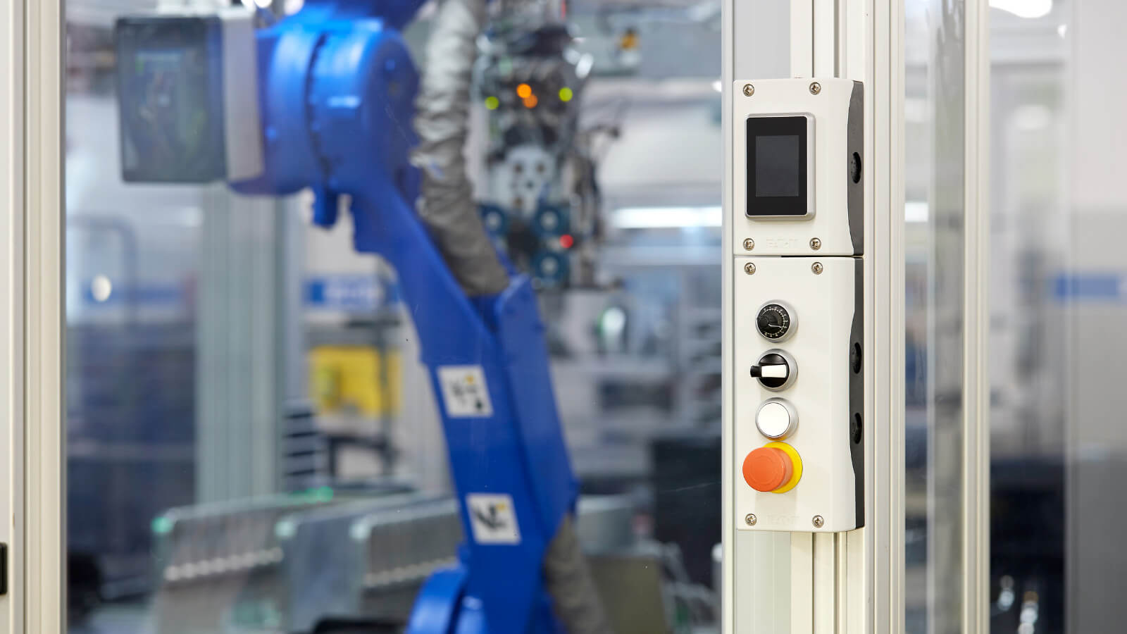 Zu sehen ist ein weißes Eaton-Bedien- und Meldeelement mit Display, Drehschalter und Not-Aus-Taster an einer Industrieanlage. Im Hintergrund erkennt man einen Roboterarm in einer Fertigungszelle.