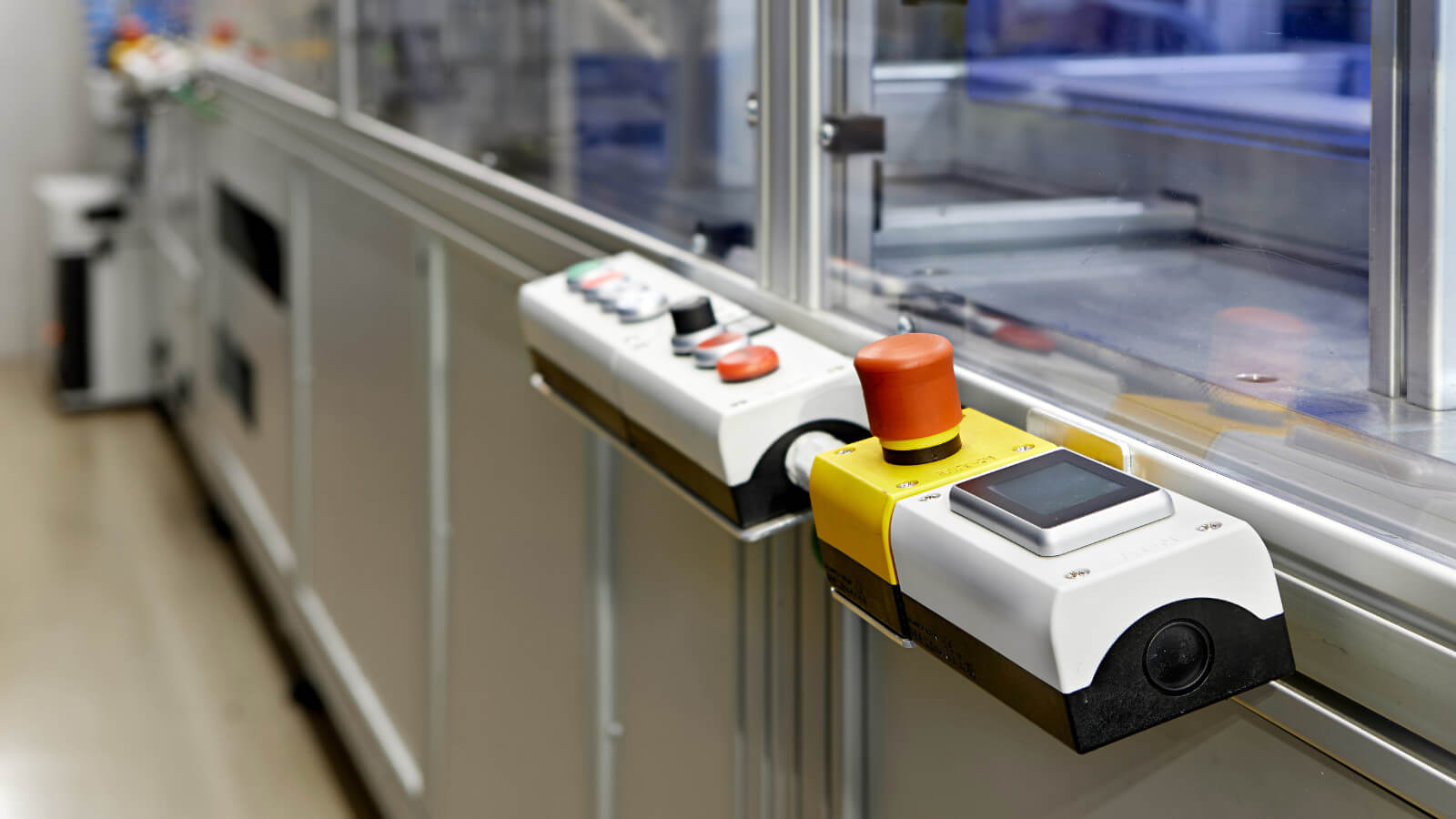 Das Bild zeigt ein Bedienfeld einer industriellen Maschine mit einem großen roten Not-Aus-Schalter. Im Hintergrund ist eine Produktions- oder Automatisierungsanlage zu sehen.