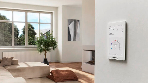 Das Bild zeigt einen hellen Wohnraum mit großem Fenster, Sofa, Topfpflanze und gemütlichen Kissen auf dem Boden. An einer Wand ist ein weißes Touch-Bedienpanel mit GIRA-Logo zu sehen, dessen Display eine Heizungssteuerung anzeigt.