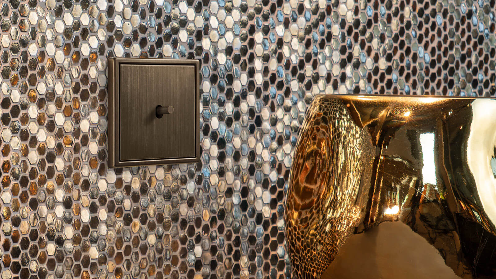 Ein Kippschalter in Bronzeoptik ist vor einer Wand mit Mosaikfliesen und neben einer goldenen Vase montiert.