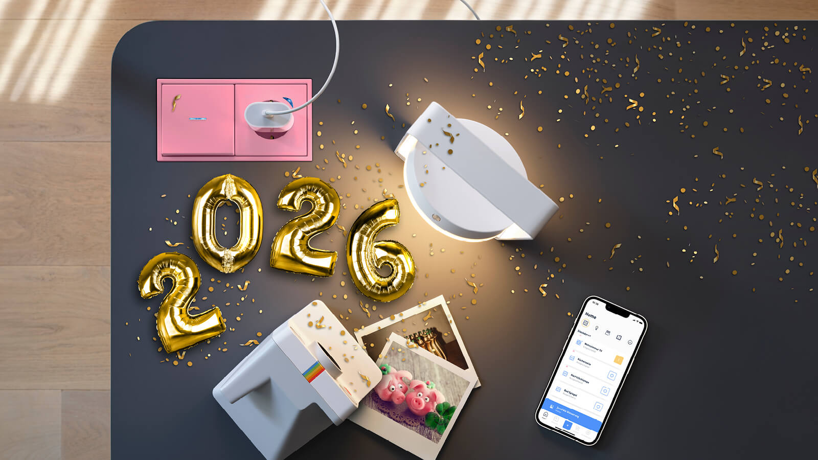 Das Bild zeigt eine Draufsicht auf eine dunkle Tischfläche mit goldenen Luftballons in Form der Zahl „2026“, verstreutem Konfetti, einer rosa Steckdose mit eingestecktem Stecker, einer weißen Leuchte, einer kleinen Fotokamera mit ausgedruckten Fotos sowie einem Smartphone mit geöffneter App.