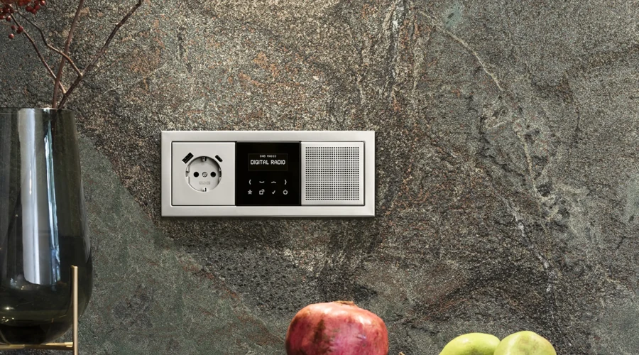 Flaches Radio zum Einbauen mit Steckdose direkt an der grauen Wand montiert. Davor liegen ein Granatapfel und eine Banane.