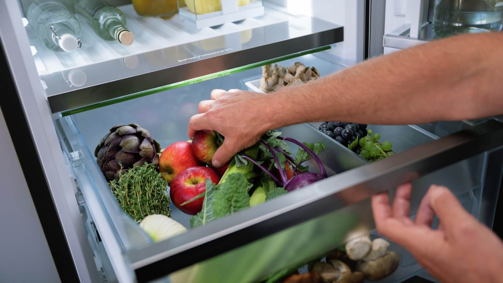Eine Hand greift in das Gemüsefach eines Kühlschranks, das mit frischem Obst und Gemüse wie Äpfeln, Artischocken, Trauben und Kräutern gefüllt ist.