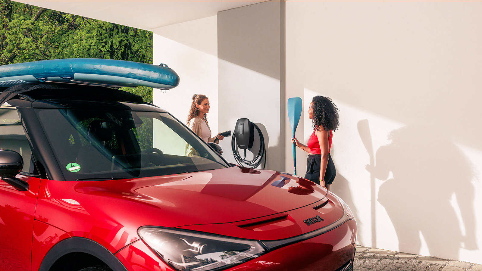 Ein roter Kleinwagen mit einem stand up paddle board auf dem Dach steht in einer offenen Garage, daneben ist eine Wallbox an der Wand montiert. Eine Person hält das Ladegerät der Wallbox in der Hand, eine andere Person ein Paddel. 