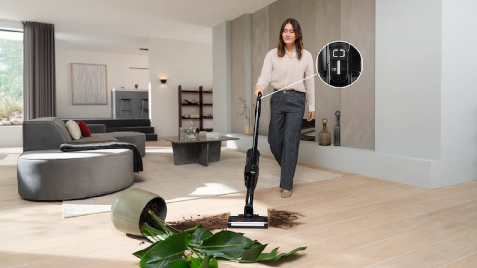 Eine Frau saugt mit einem schwarzen Stielstaubsauger Erde und Pflanzenreste auf einem hellen Holzboden auf. Ein umgekippter Blumentopf und eine Pflanze liegen daneben. Im Hintergrund ist ein modernes Wohnzimmer mit einem grauen Sofa und einem Couchtisch zu sehen. Ein Detailausschnitt zeigt ein Display am Staubsauger.