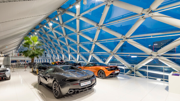 Das Bild zeigt einen modernen Ausstellungsraum mit mehreren Sportwagen. Die Halle hat eine große Glasfassade mit diagonalen Stahlträgern.