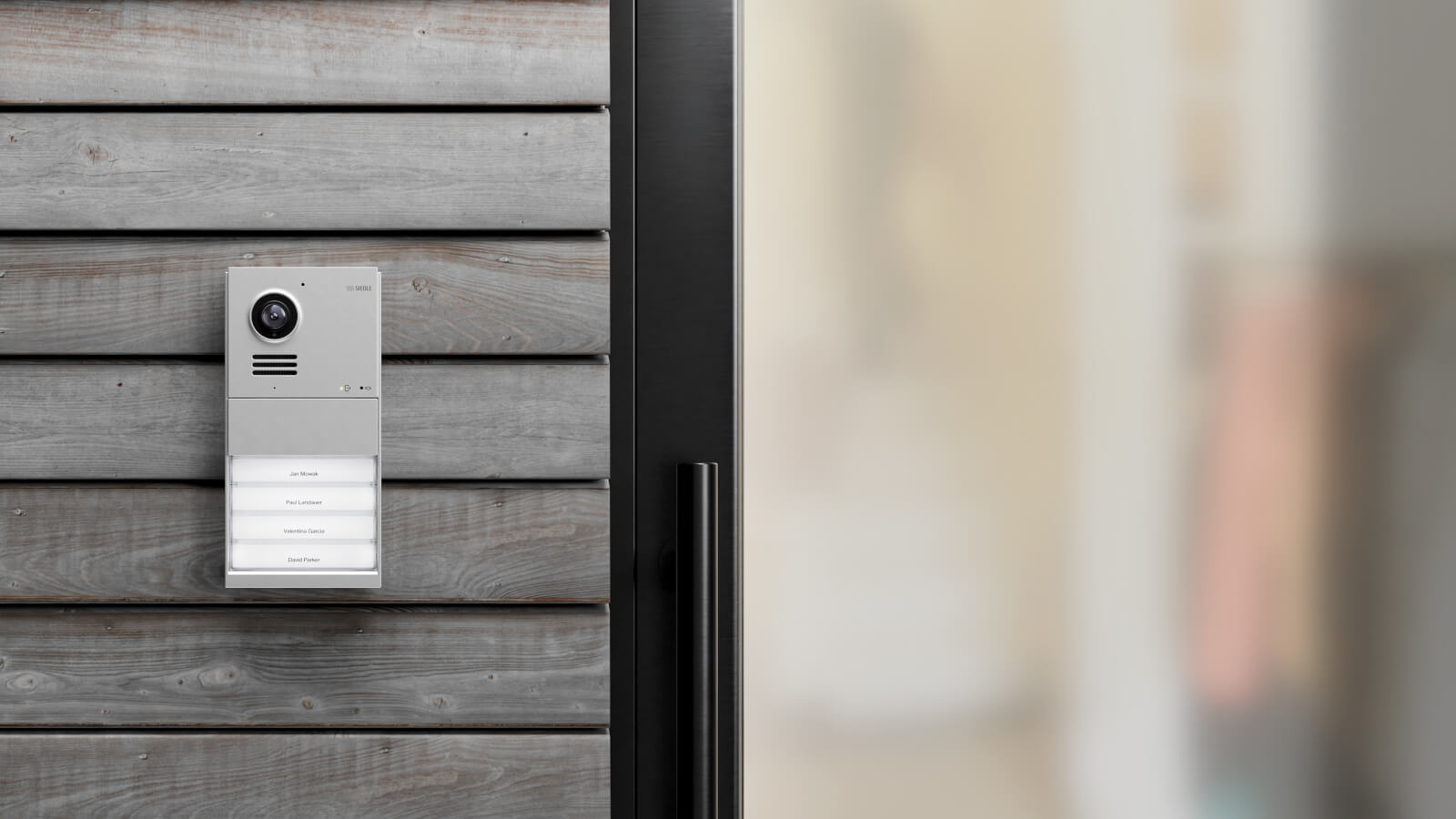 Das Bild zeigt eine graue Siedle IQ Außenstation an einer grauen Holzwand. neben einer schwarzen Haustür.