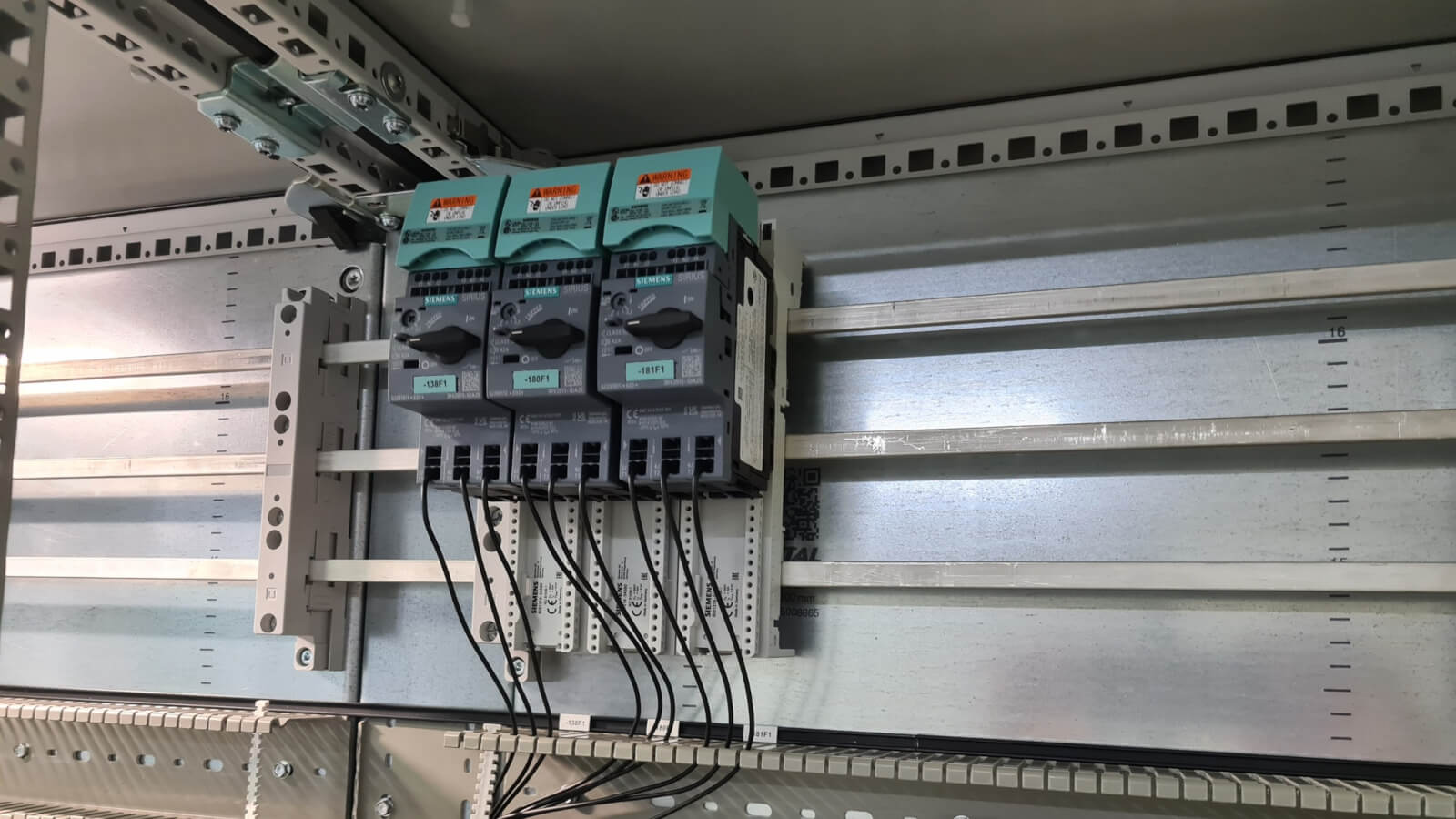 Einspeisesystem 3RV29 Das Bild zeigt einen Ausschnitt eines Schaltschrankes. Darin sind drei Schaltgeräte des Typs „SIEMENS SIRIUS“ montiert. Jedes Gerät ist mit einem Drehschalter versehen und mit einer grünen Beschriftung versehen (z. B. „-13BF1“, „-18DF1“). Unterhalb der Geräte sind schwarze Kabel angeschlossen, die nach unten in einen Kabelkanal geführt werden. Die Geräte sind auf einer Metallmontageschiene befestigt. Links neben den Schaltgeräten ist ein weiteres, leeres Trägerelement montiert. Im Hintergrund ist eine Metallrückwand mit Maßskala zu sehen.
