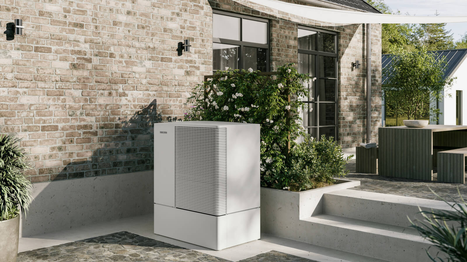 Das Bild zeigt eine moderne, weiße Wärmepumpe von Stiebel Eltron, die auf einer Terrasse vor einer Hauswand mit Klinkersteinen installiert ist. Die Umgebung ist grün und gepflegt.