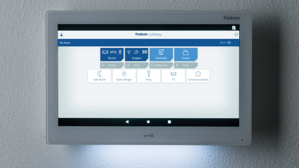 LUXORliving CP10: Innenstation für smarte Türkommunikation und zentrale Smart Home-Steuerung Das Bild zeigt ein weißes LUXORplay-Display, das an der Wand montiert ist und eine integrierte Beleuchtung an der Unterseite des Geräts aufweist. Diese Beleuchtung sorgt für eine sanfte, zusätzliche Atmosphäre. Die Benutzeroberfläche des Displays ist identisch mit den anderen, mit Icons zur Steuerung von Funktionen wie „Gute Nacht“, „Party“ und „TV“. Auf dem Bildschirm ist eine Temperaturanzeige von 25°C sichtbar.