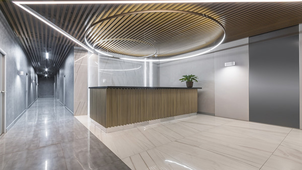 TRILUX OSIDO Das Bild zeigt eine moderne und stilvolle Empfangshalle. Im Zentrum befindet sich ein elegant gestalteter Empfangstresen mit vertikalen Holzlamellen und einer dunklen Arbeitsfläche. Darüber hängt eine auffällige, runde Lichtinstallation, die von einer Decke mit Holzlamellen ergänzt wird und dem Raum eine warme Atmosphäre verleiht. Die Wände und der Boden sind in hellen, glatten Materialien gehalten, teilweise mit Marmoroptik. Links und rechts führen Korridore in weitere Bereiche des Gebäudes. Auf dem Tresen steht ein grüner Topfpflanze, die einen frischen Akzent setzt.