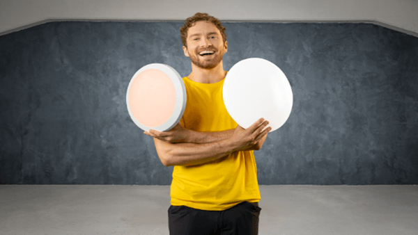 TRILUX twenty3 Ein Mann in gelbem T-Shirt und schwarzer Hose steht in einem Raum mit grauer Wand. Er hält in jeder Hand eine runde Leuchte – eine mit warmem Licht, die andere ohne sichtbares Licht. Er schaut in die Kamera und lächelt.