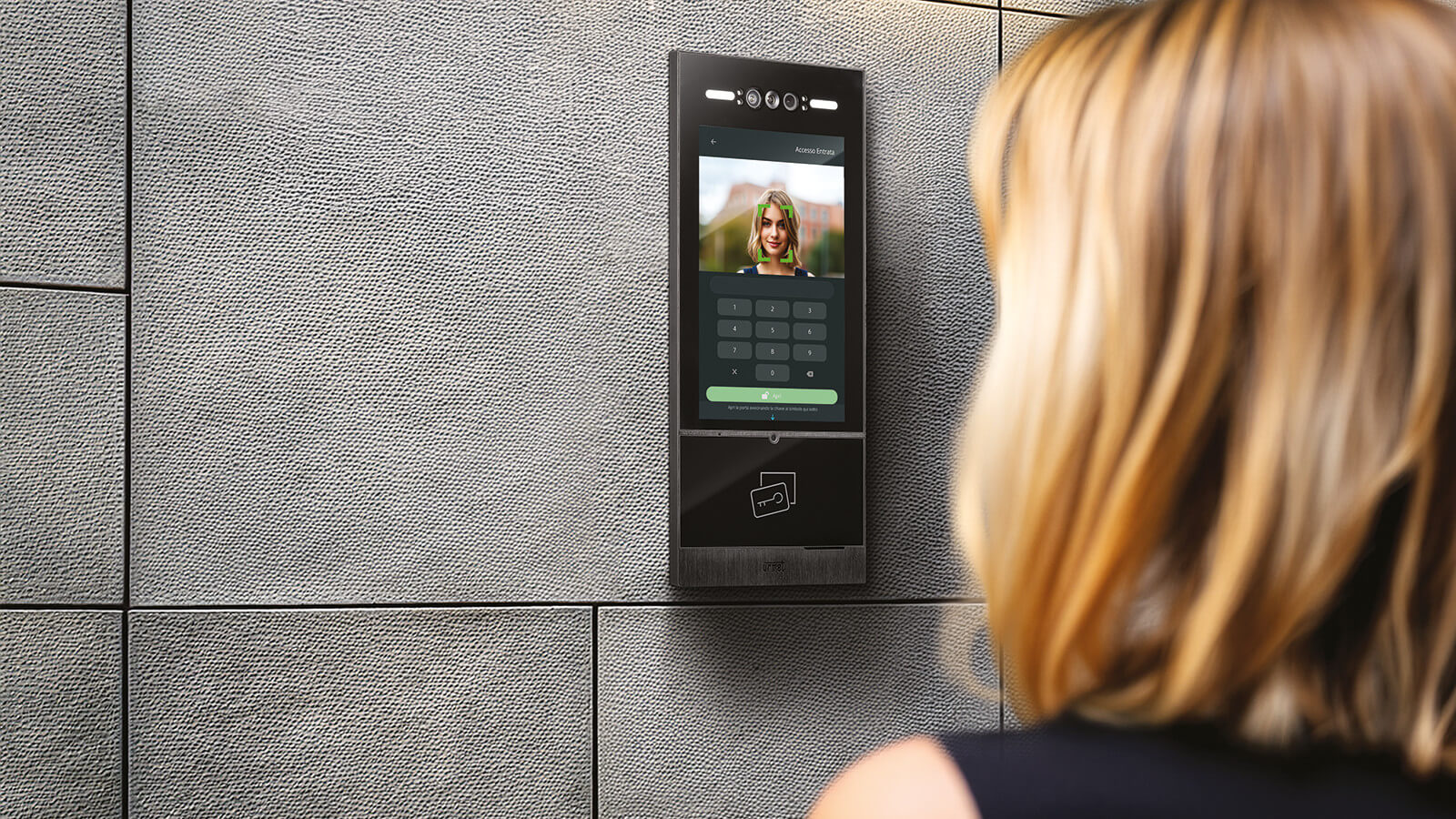 Eine Person steht vor einer wandmontierten Türstation mit Touchdisplay. Auf dem Bildschirm ist ein Porträtfoto mit grünen Markierungen im Gesichtsbereich zu sehen.