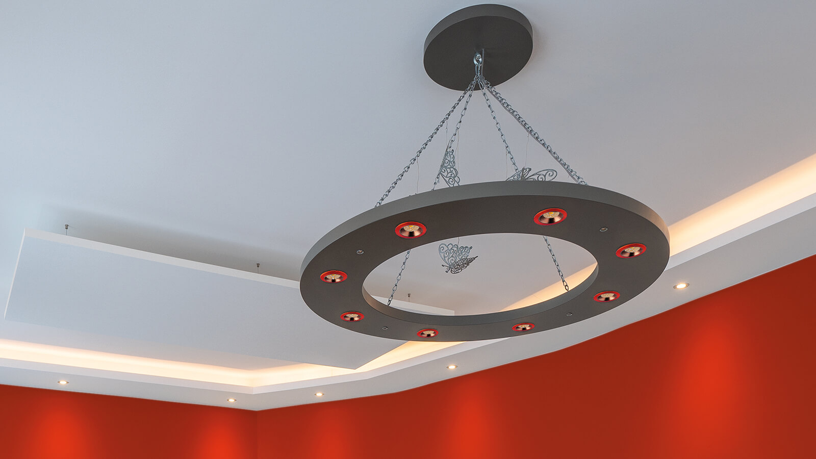 Zu sehen ist eine moderne, ringförmige Deckenleuchte, die an Ketten von der Decke hängt. Die Szene zeigt einen Innenraum mit roten Wänden und indirekter Beleuchtung.
