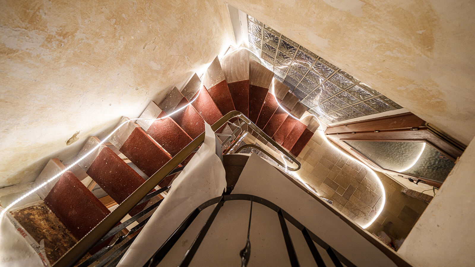 Das Bild zeigt eine von oben fotografierte Wendeltreppe im Rohbau, die von einer LED-Lichtleiste beleuchtet wird. Die Stufen sind mit rotem Teppich belegt und die Wände sind unverputzt.