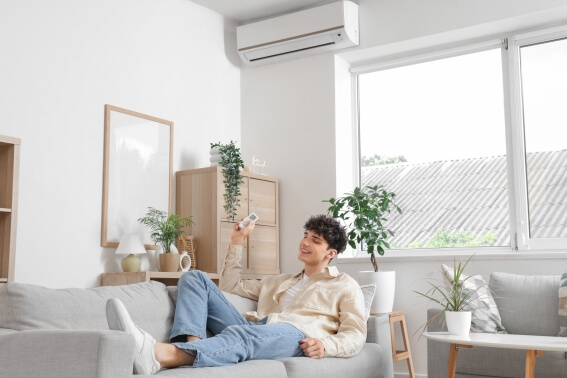 Person sitzt gemütlich auf dem Sofa und hat eine Fernbedienung in der Hand mit der er die Klimaanlage im Hintergrund steuert.