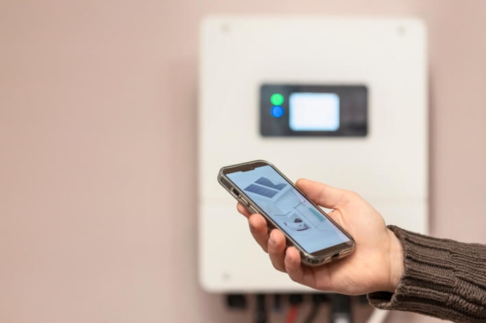 Hand mit Smartphone, dahinter ein Stromspeicher an der Wand