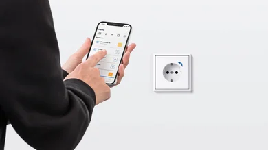 Person hält ein Smartphone in der Hand und bedient das Display. Aufnahme von leicht schräg hinten. An einer weißen Wand ist eine weiße Steckdose angebracht.
