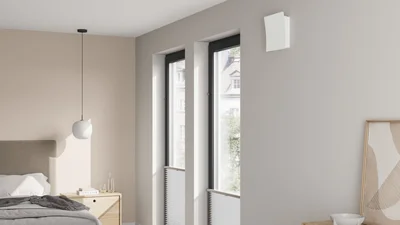 Heller Schlafraum mit Bett, Nachttisch und zwei hohen Fenstern. Oberhalb der Fenster ist ein kompaktes, weißes Lüftungsgerät montiert.