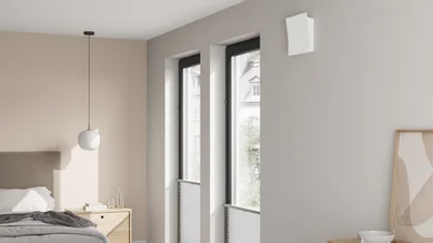 Heller Schlafraum mit Bett, Nachttisch und zwei hohen Fenstern. Oberhalb der Fenster ist ein kompaktes, weißes Lüftungsgerät montiert.