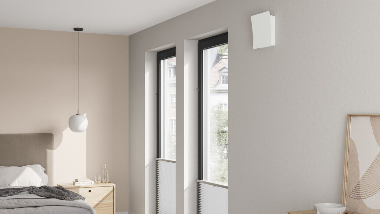 Heller Schlafraum mit Bett, Nachttisch und zwei hohen Fenstern. Oberhalb der Fenster ist ein kompaktes, weißes Lüftungsgerät montiert.