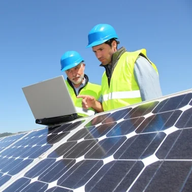 Das Bild zeigt zwei Personen mit Schutzhelm und Warnweste, die hinter einer Photovoltaikanlage stehen und etwas am Laptop begutachten.