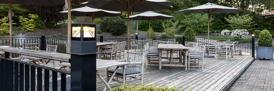 Das Bild zeigt eine Terrasse mit Holzbohlen. Auf der Terrasse stehen Holztische in verschiedenen Größen, an den Stühle stehen. Um die Terrasse herum verläuft ein Metallgeländer. Oben auf den Geländerpfosten sind Leuchten integriert. Eine die Leuchten befindet sich im Vordergrund des Bildes.