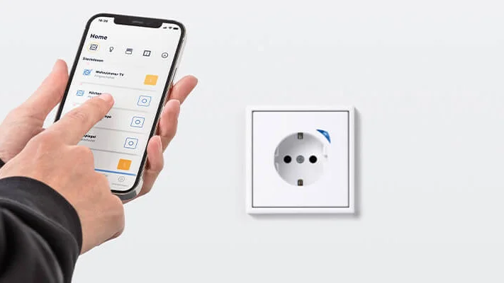 Eine Person hält ein Smartphone in der Hand und bedient das Display. Aufnahme von leicht schräg hinten. An einer weißen Wand ist eine weiße Steckdose angebracht.