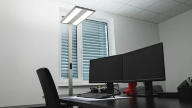 Das Bild zeigt einen schwarzen Schreibtisch mit Stuhl und zwei Bildschirmen. Neben dem Schreibtisch steht vor einem Fenster mit geschlossenem Rollladen eine Büroleuchte MASTERhcl von Dotlux.