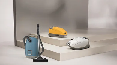 Drei verschiedene Ausführungen des Guard Staubsaugers von Miele in den Farben Blau (vorne) Weiß und Organge. Vor einem grauen Hintergrund.