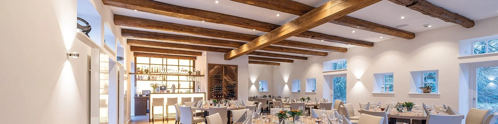 Zu sehen ist ein hell eingerichteter Restaurant- oder Gastraum mit Holzbalkendecke, gedeckten Tischen und einer Bar im Hintergrund. Die warmen Wand- und Deckenleuchten sorgen für eine gemütliche Atmosphäre.