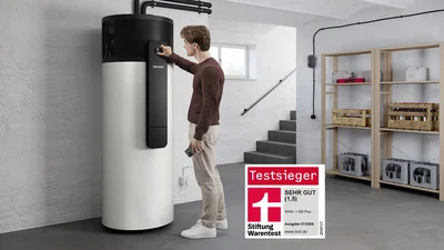 Das Bild zeigt eine Person, die eine Wärmepumpe in einem Keller- bzw. Hauswirtschaftsraum bedient. Rechts stehen Regale mit Batteriemodulen und Kisten. Unten rechts ist ein „Testsieger“-Siegel der Stiftung Warentest für das Modell WWK-I 300 Plus zu sehen