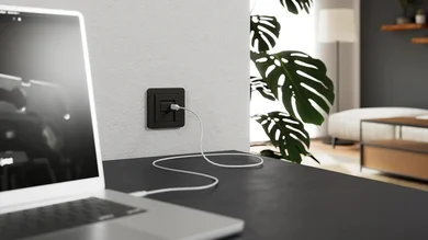 Das Bild zeigt ein Notebook auf einem dunklen Tisch, das über ein weißes Kabel an eine schwarze USB-Steckdose an der Wand angeschlossen ist. Im Hintergrund ist eine große Monstera-Pflanze und ein Wohnbereich erkennbar.