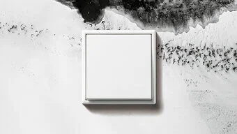 Das Bild zeigt einen Schalter des Herstellers Jung in der Farbe Mattweiß, der an einer schwarzweiss gemusterten Wand befestigt ist.