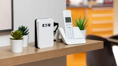 Auf einem Regalboden aus Holz steht eine Eaton 3S Mini USV neben einem weißen, schnurlosen Telefon. Links und rechts von Eaton 3S Mini USV und Telefon stehen kleine Pflanzen in weißen Übertöpfen. Im Hintergrund ist ein Schubladenschrank in orange erkennbar.