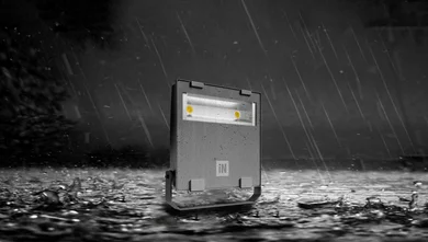 Das Bild zeigt einen grauen LED-Strahler mit zwei Lichtpunkten und „iN“-Logo, stehend auf nassem Boden bei starkem Regen. Der Strahler ist mit Wassertropfen bedeckt, der Hintergrund ist dunkel.