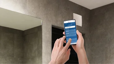 Zwei Hände halten ein Smartphone in Richtung einer oben an einer Wand befestigten Lüftung. Auf dem Display des Smartphones ist die MAICO@NFC-App erkennbar. 
