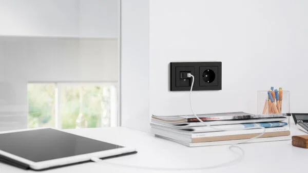Das Bild zeigt eine weiße Arbeitsfläche im Detail mit Zeitschriftenstapel, Stiftebox und Taschenrechner. Ein weißes Tablet ist über ein Ladekabel mit einer schwarzen USB-Steckdose verbunden, die an einer weißen Wand installiert ist. 