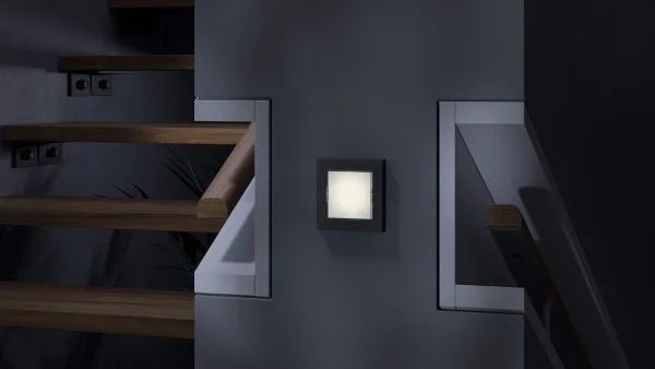 Das Bild zeigt den Teil eines modernen Treppenhauses mit Holztreppe, das im Dunkeln liegt. An einer Wand ist ein Orientierungslicht befestigt, das leuchtet.