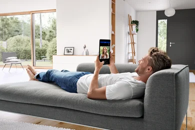 Das Bild zeigt eine Person auf einem Sofa, die das Bild einer anderen Person auf einem Tablet betrachtet. 