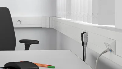 Helles Büro mit weißem Schreibtisch und schwarzem Bürostuhl sowie Geräteeinbaukanal von OBO Bettermann rechts an der Wand.